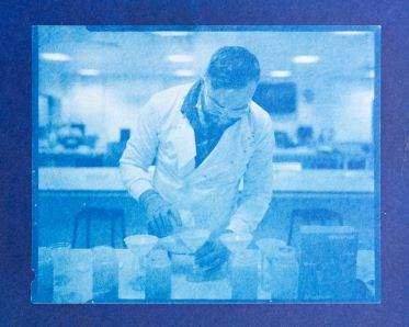 A bright blue cyanotype image with a dark blue frame showing a man wearing a white lab coat in a laboratory setting, pouring liquid through a funnel into a beaker.