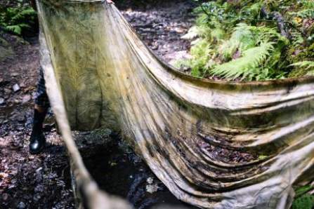 A large length of wet cotton fabric is stretched out against a backdrop of ferns, dappled light and a forest floor. The fabric is muddy, ripped and stained.