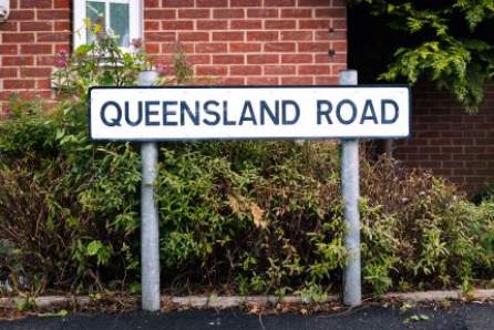 A road sign reading ‘Queensland Road’ in black lettering on a white background, attached to two metal post, in front of a green shrubby bush and part of a red brick building.