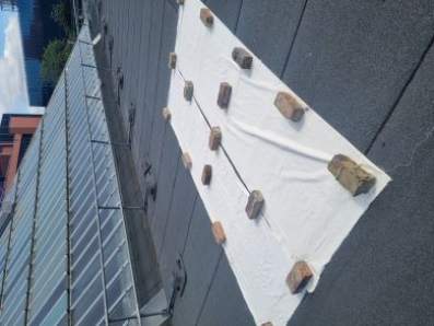 Two lengths of cotton fabric are stretched out on a flat, grey roof, weighted down with bricks.