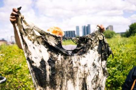 A man holds a large piece of ripped cotton fabric, marked with black mud, against a backdrop of greenery, cloudy sky, and tall buildings in the distance. His hands and the top of his head are visible.