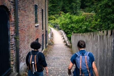 Two women wearing rucksacks pictured from behind walk down a cobbled path towards a small bridge and green trees, a large red brick mill building on their left.