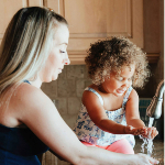 A woman helping her child wash her hands at a kitchen sink.